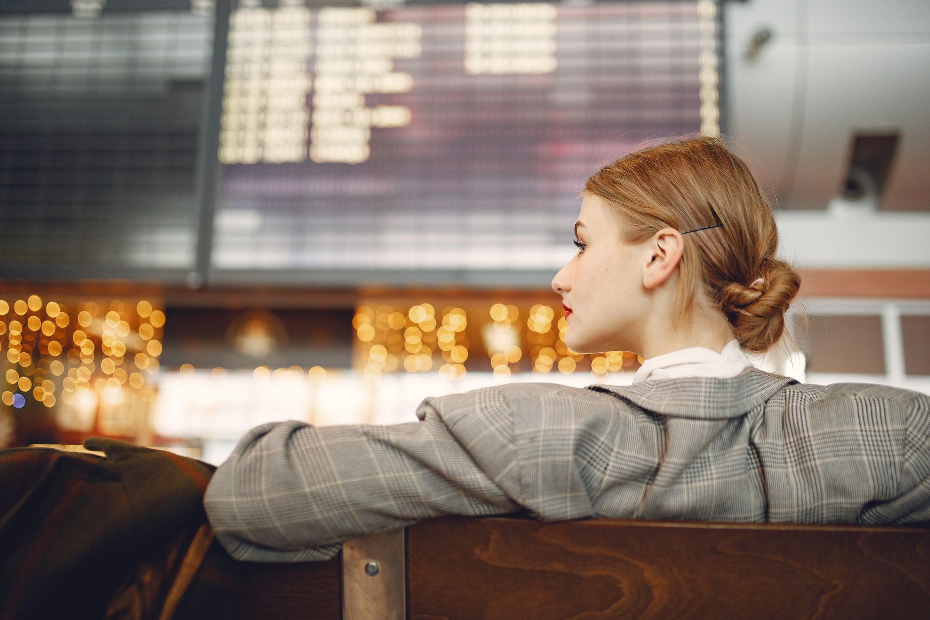 Designer féminine pensive en veste à carreaux attendant son vol près du tableau d'affichage, regardant ailleurs dans un aéroport moderne en soirée.
