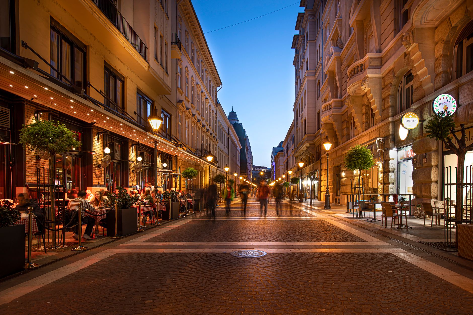 Rue piétonne pavée au crépuscule, avec terrasses de restaurant éclairées, lampadaires allumés et passants flous entre des immeubles anciens.