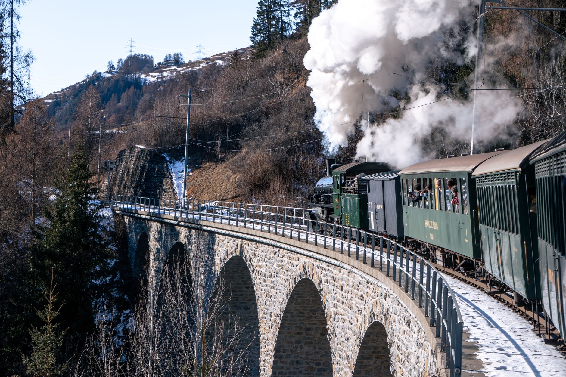 Train à vapeur ancien aux voitures vertes sur un viaduc en pierre en courbe, en montagne enneigée, avec un épais panache de vapeur.